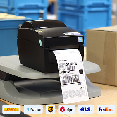 Versandetikettendrucker mit ausgedrucktem Label für UPS auf einem Packtisch, darunter Logos von geeigneten Versanddienstleister.
